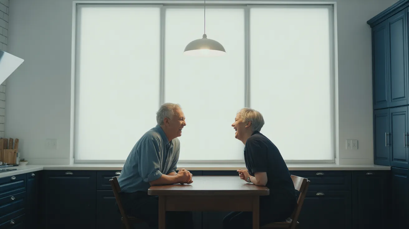 An elderly man and woman share a laugh over coffee at a modern kitchen table, seen in a symmetrical, brightly lit medium shot.