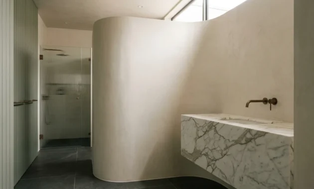 A luxury bathroom featuring seamless bone-colored plaster walls, a large marble vanity, and dark floor slabs under soft natural light.