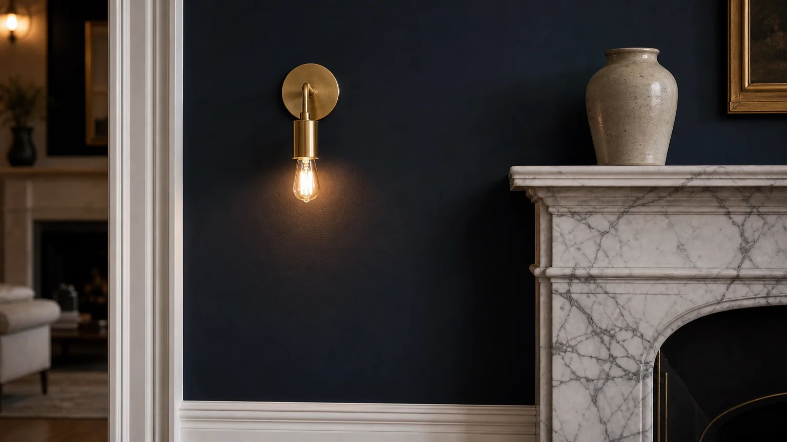 A close-up of a living room corner with matte dusty navy walls, glossy white crown molding, and an unlacquered brass sconce.