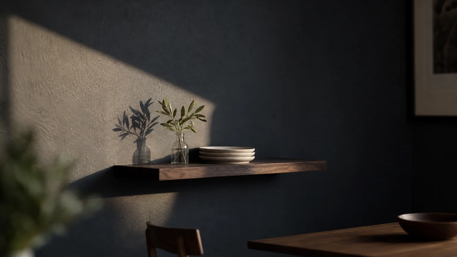 A close-up of a matte charcoal black wall with a dramatic beam of sunlight, a dark wood shelf, white plates, and a green sprig.