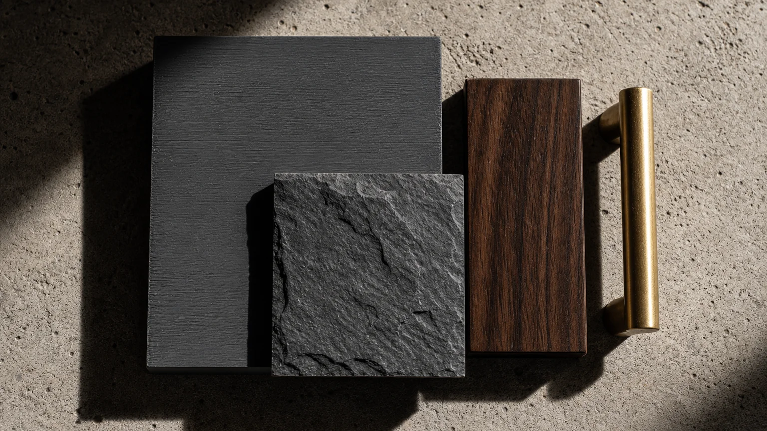 A flat lay material board featuring a soft charcoal paint swatch, dark slate, walnut wood, and a brushed brass pull.