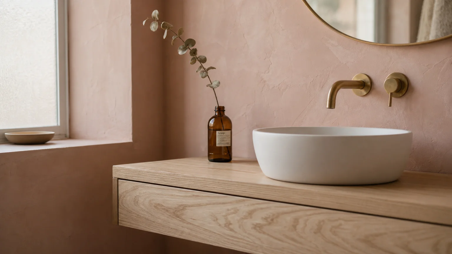 A minimalist bathroom corner with a dusty plaster pink wall, a light ash vanity, a white vessel sink, and a brass faucet.