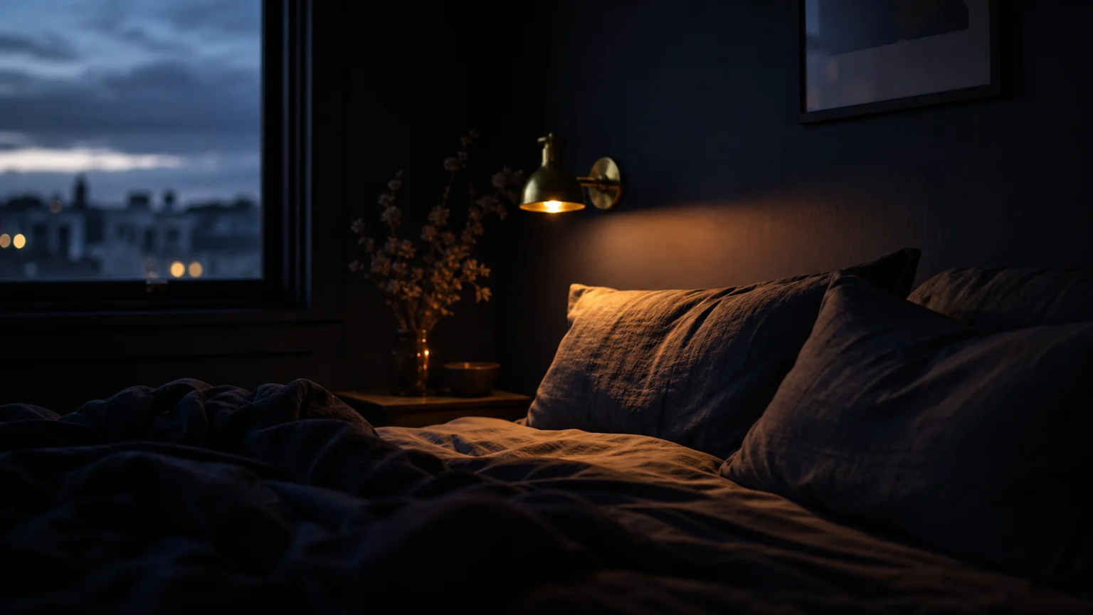 A moody, dark navy bedroom at twilight, illuminated by the warm, soft glow of a single brass wall sconce over dark linen bedding.