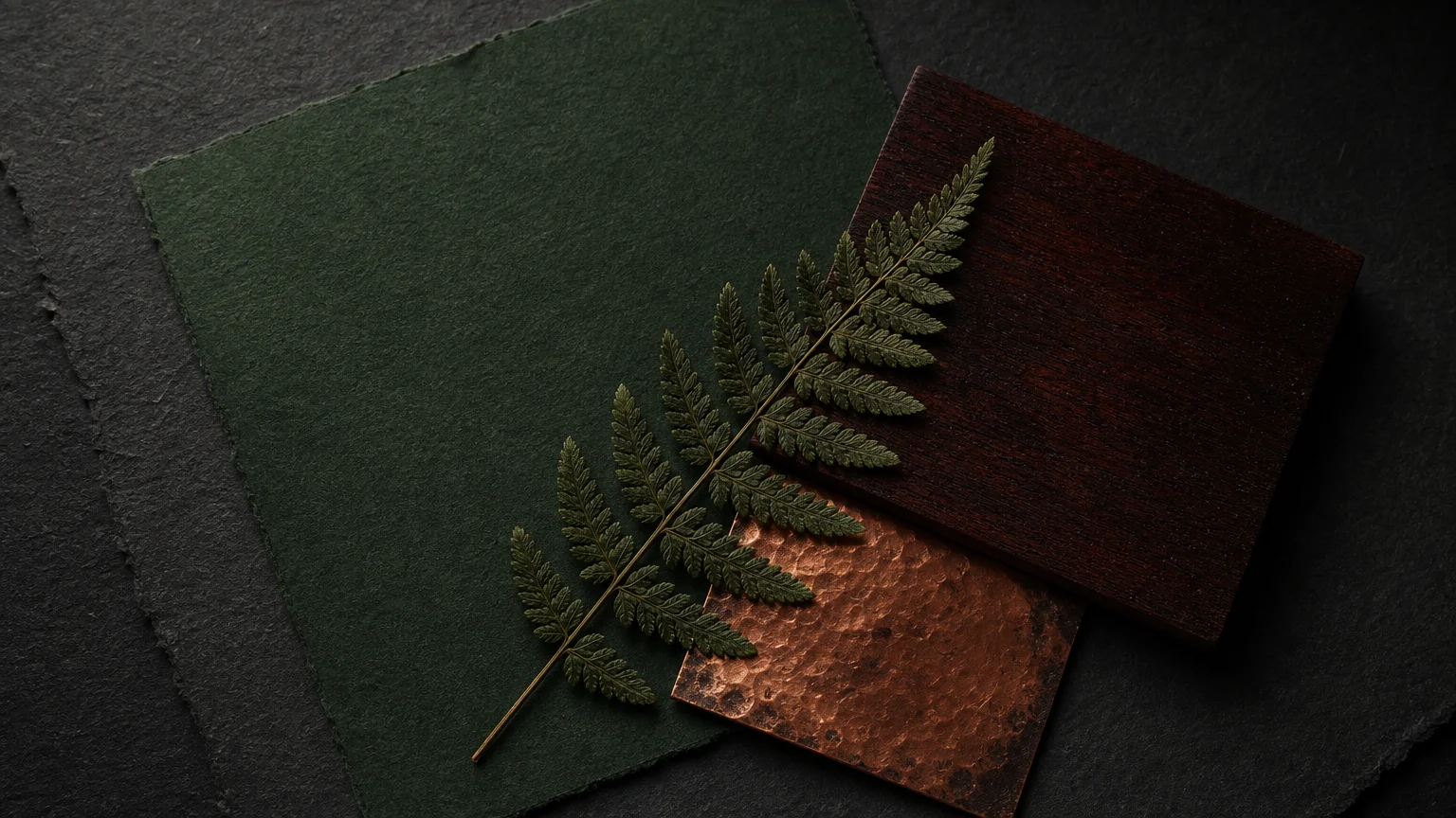 A moody mood board with a deep forest green paint swatch, dark mahogany wood, tarnished copper, and a pressed fern.