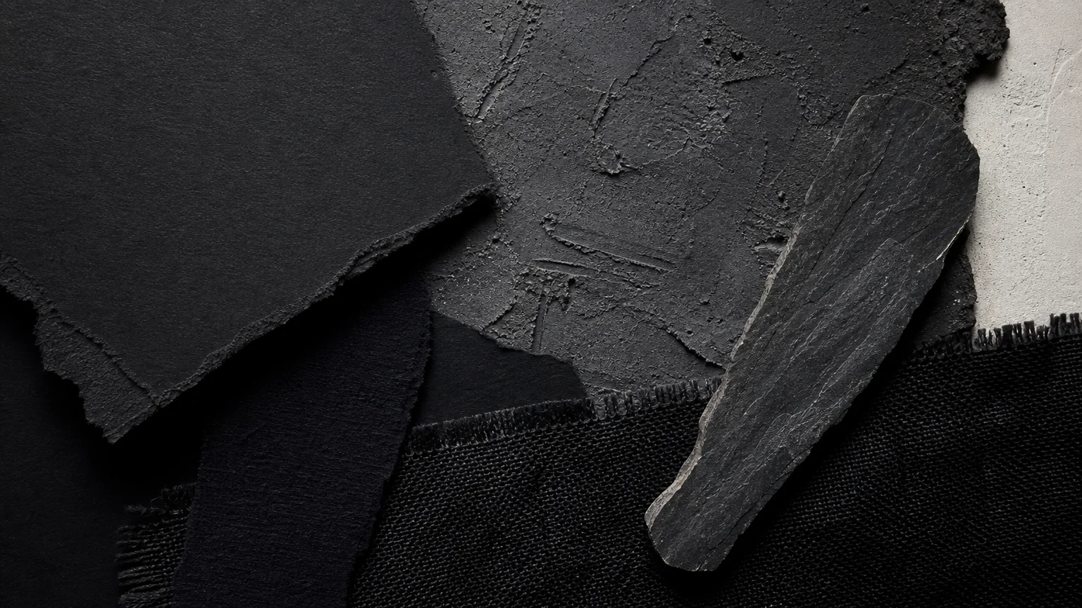 A tactile collage of overlapping charcoal-grey cardstock, black linen fabric, and natural dark slate.