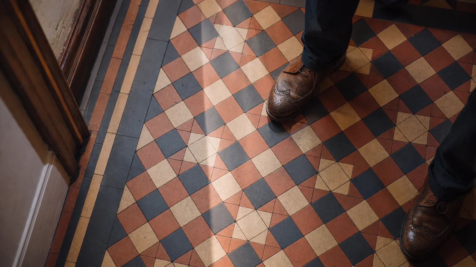 A top-down photograph of geometric encaustic floor tiles in terracotta and charcoal, with leather shoes walking on them.