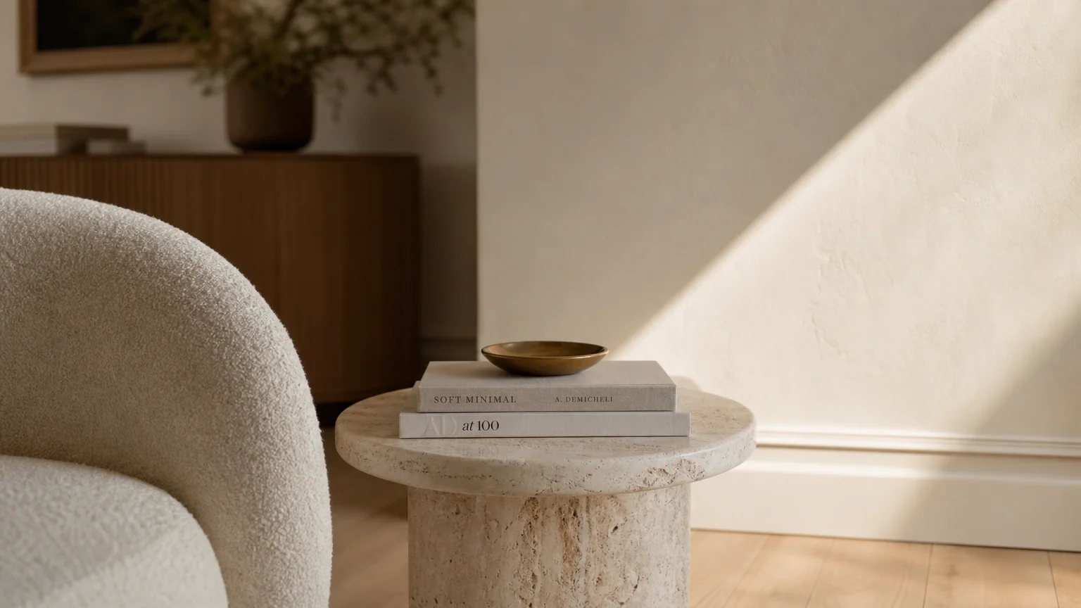 Close-up of a warm alabaster white wall in soft sunlight, next to a cream bouclé chair and a travertine stone side table.