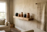 A modern living room wall displaying a white oak floating shelf with ceramic urns and a linen tapestry under warm afternoon light.