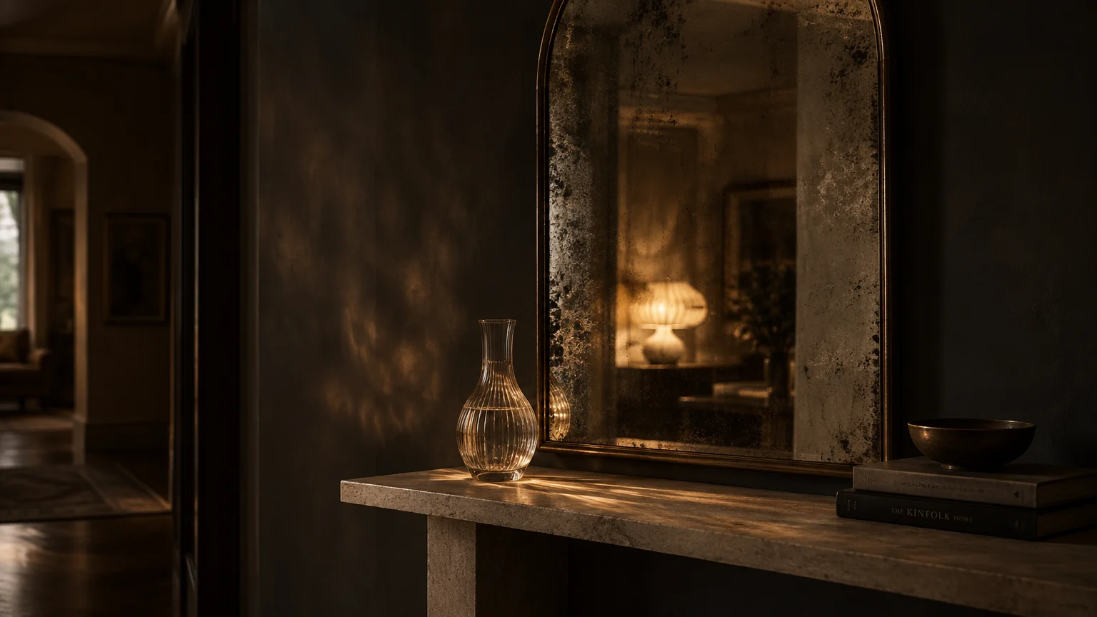Antiqued arched mirror on a stone console table reflecting a glowing lamp beside a glass carafe and books.