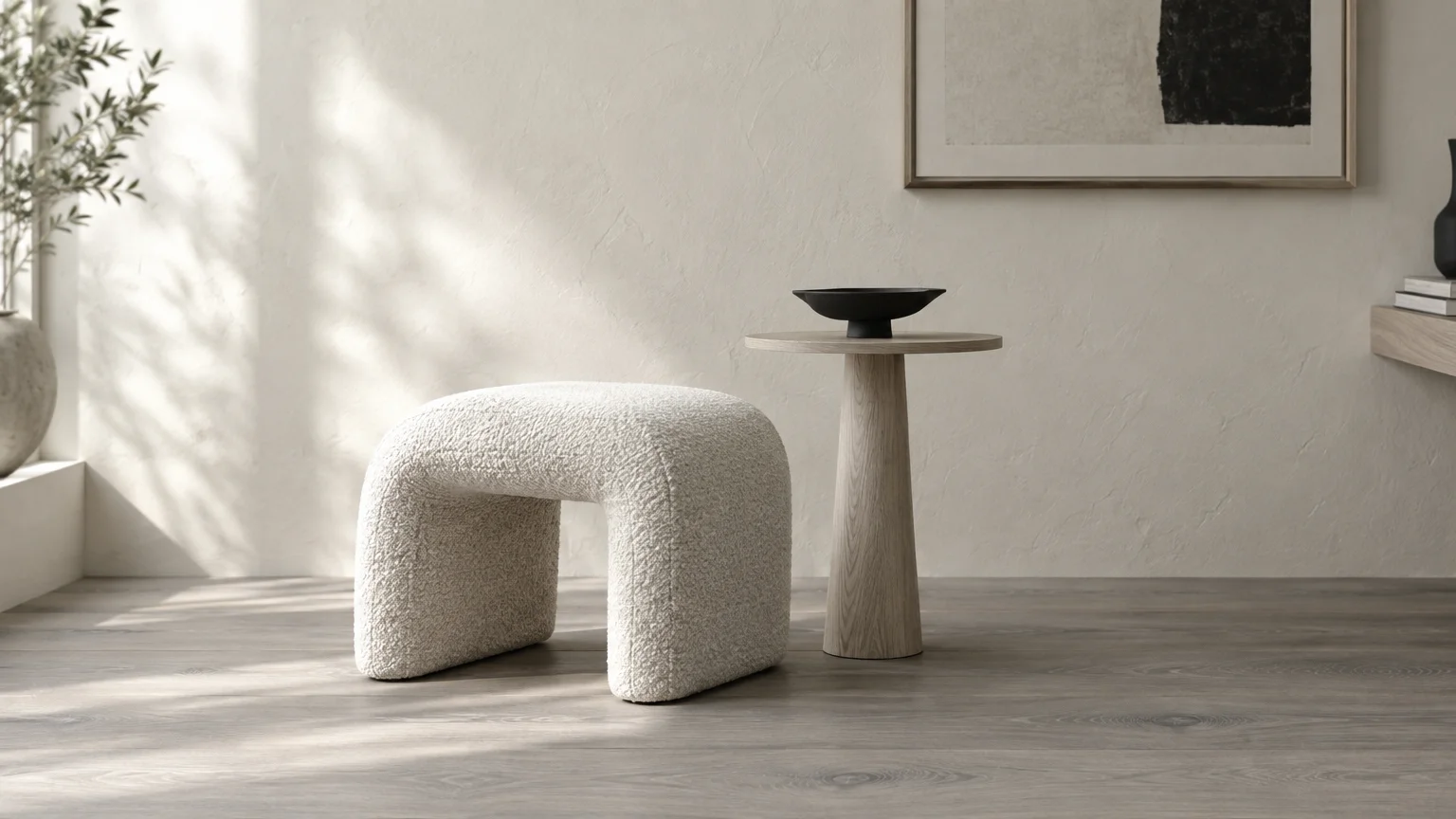 White bouclé arched stool beside a round wood pedestal table with a black bowl on cool-toned wood floors.