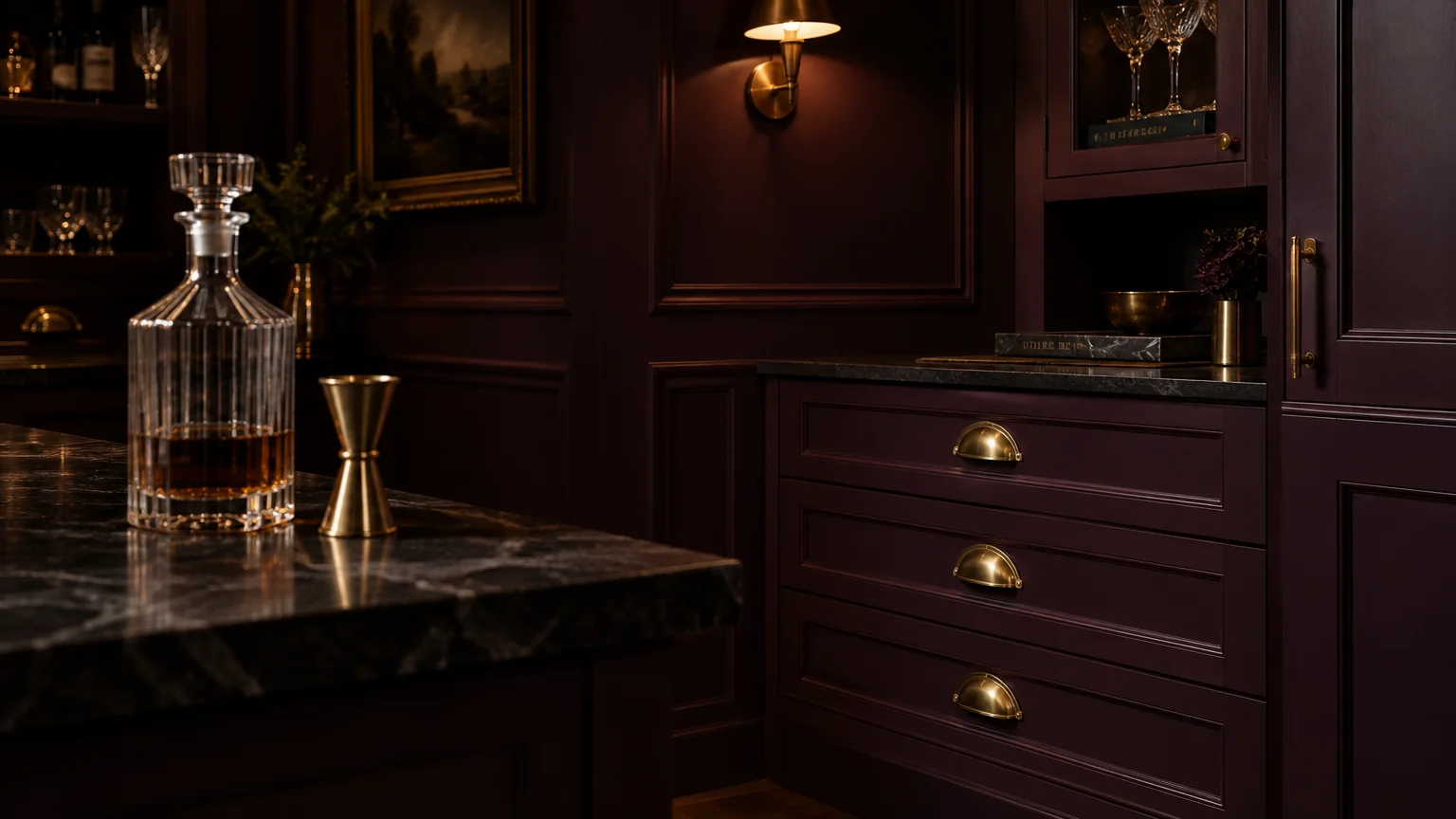 Dark plum cabinetry with brass cup pulls, dark marble countertops, a crystal decanter, and a brass wall sconce.