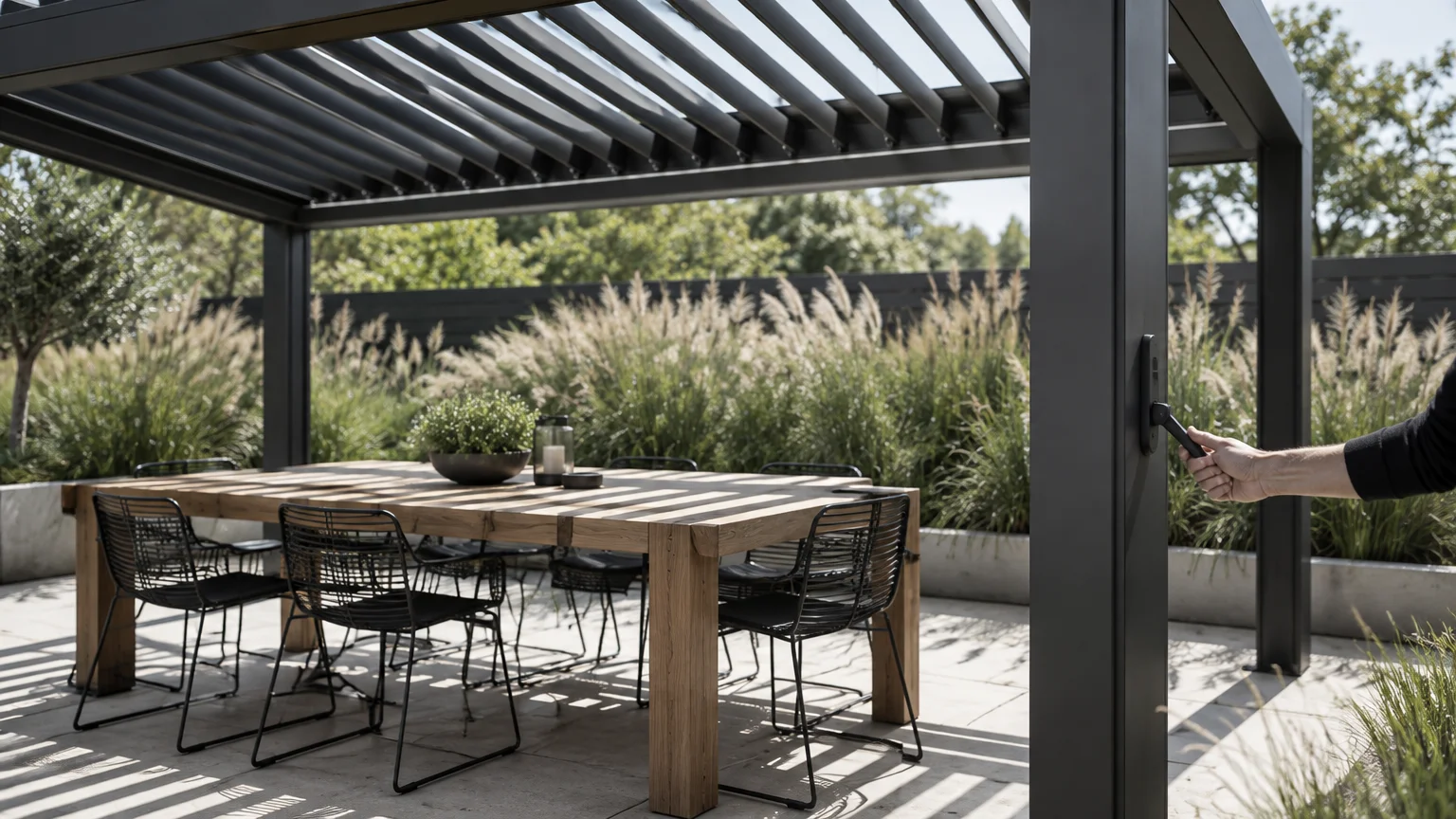 A black aluminum louvered pergola over a wooden dining table and black chairs on a patio with tall ornamental grasses.