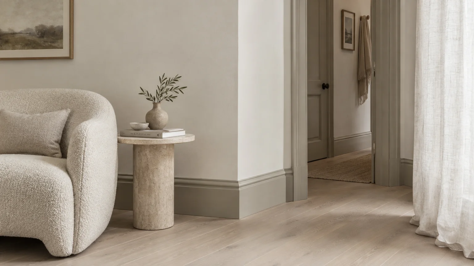 Off-white living room featuring a boucle armchair, stone side table, greige baseboards, and a doorway leading to a hallway.