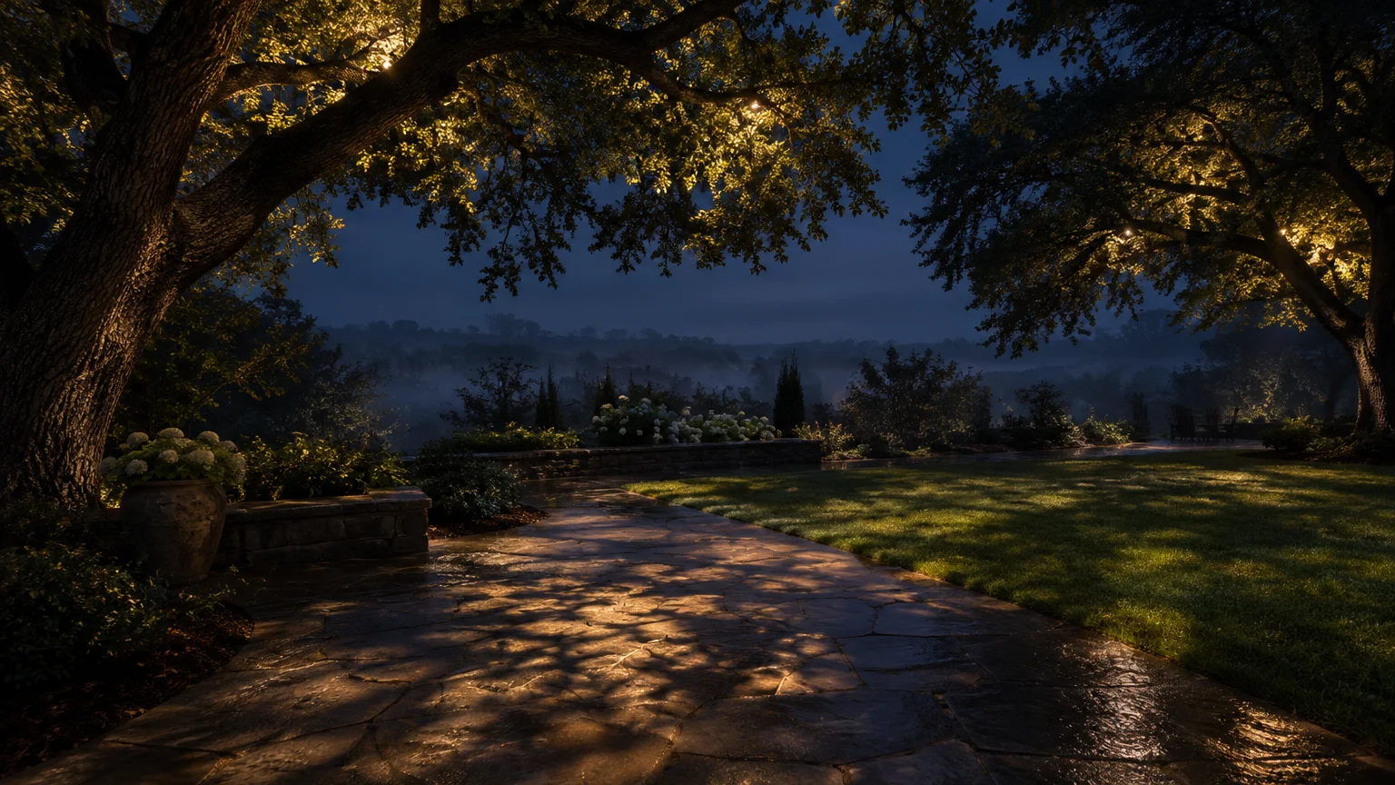 Downward warm lights mounted in mature trees cast dappled shadows over a stone patio and lawn at twilight.