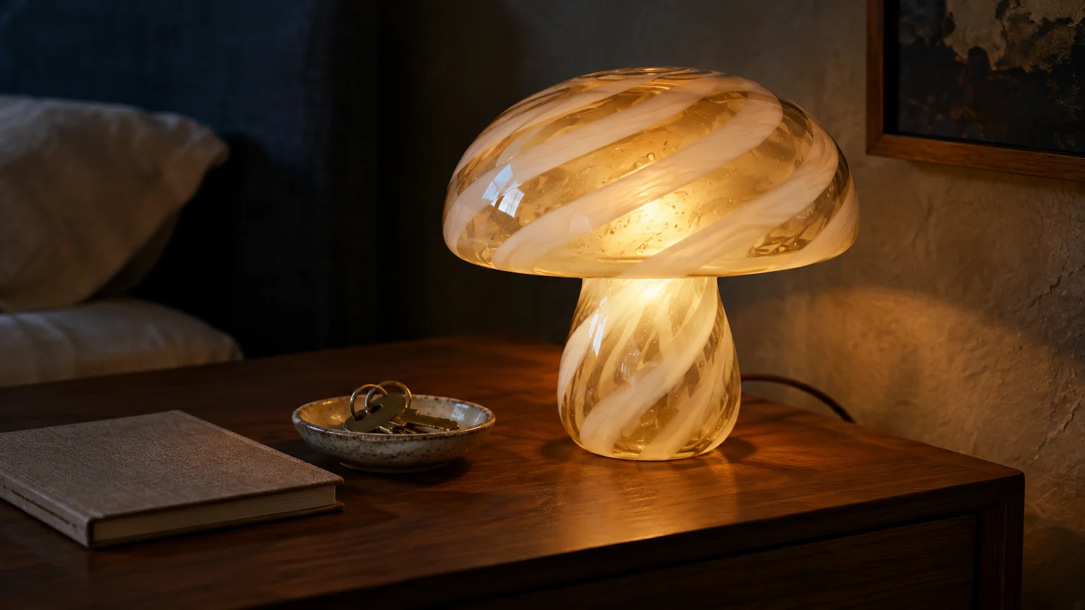 Illuminated glass mushroom lamp with white swirls glowing warmly on a wooden nightstand beside a book and key dish.