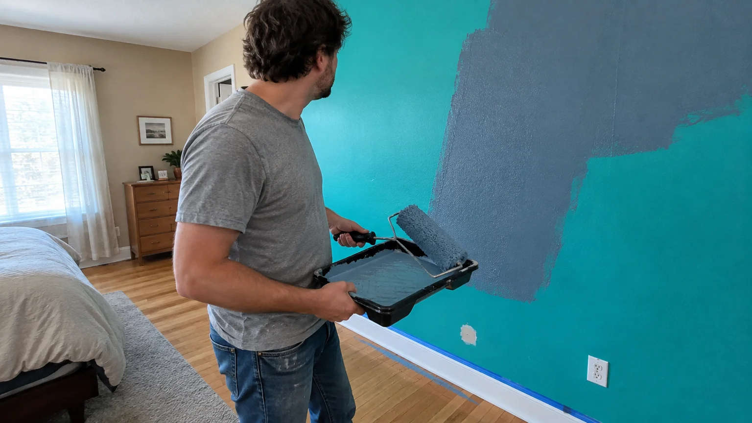 A man rolls muted gray paint over a vibrant electric teal bedroom accent wall with a paint roller and tray.