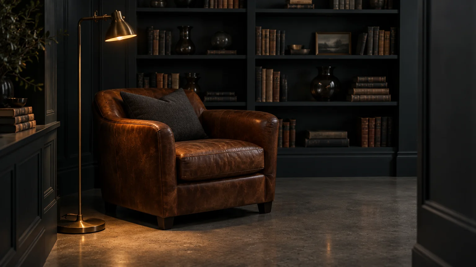 Brown leather armchair next to a brass floor lamp on a concrete floor in a dark charcoal room with built-in bookshelves.