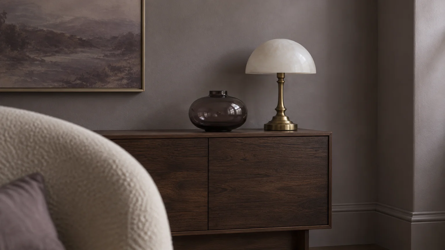 A dome lamp, dark glass vase, and framed landscape painting on a wooden sideboard against a muted greige wall.