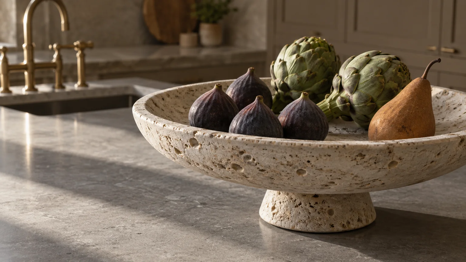 Footed travertine bowl on a stone island holding fresh figs, green artichokes, and a pear in natural sunlight.