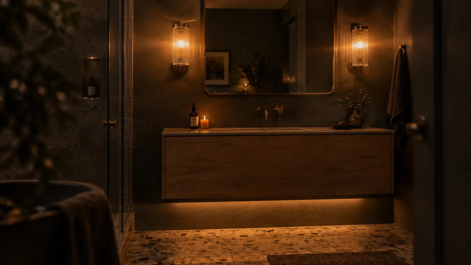 Warmly lit modern bathroom vanity with two fluted glass wall sconces, under-cabinet lighting, and terrazzo floor tiles.