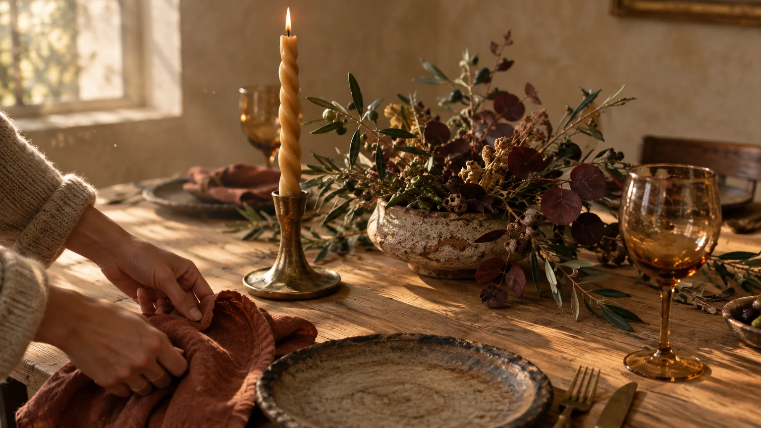 Hands arranging a rust cloth napkin beside a ceramic plate, twisted candle in brass holder, and rustic floral centerpiece.