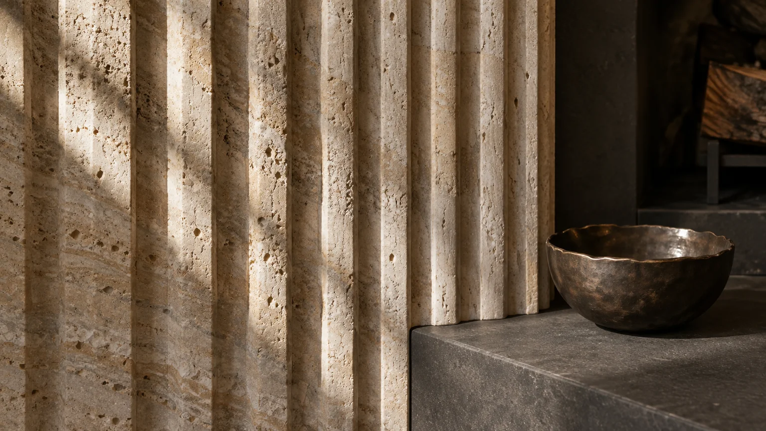 Fluted beige travertine fireplace surround beside a dark stone hearth holding a small hammered bronze bowl.