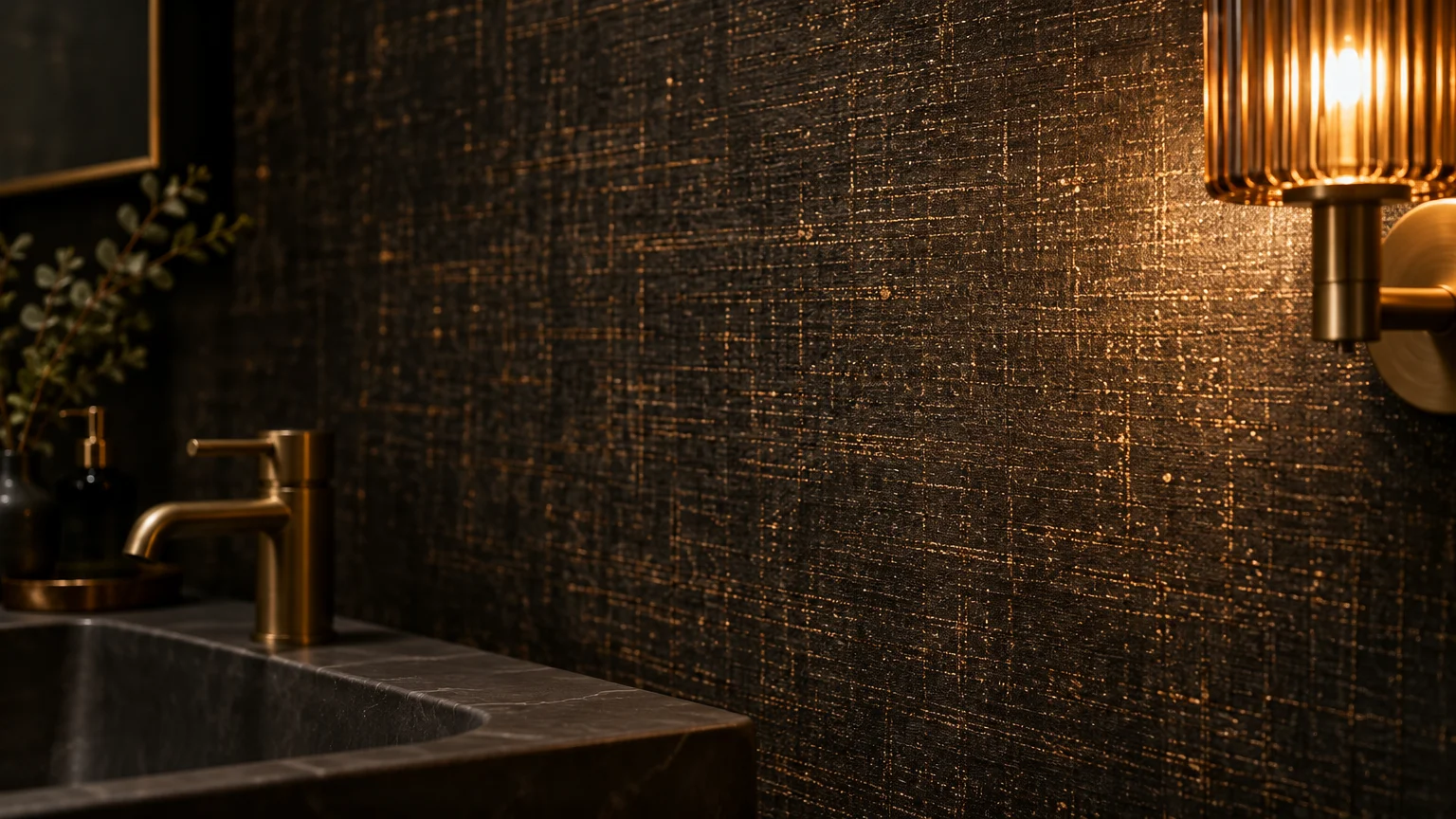 Dark textured metallic wallpaper illuminated by a fluted brass wall sconce beside a black marble sink.