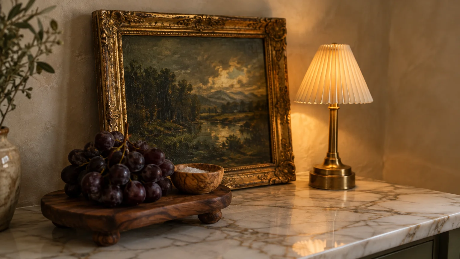 Framed oil landscape painting leaning on a marble counter beside a glowing small lamp, footed wooden trivet, and grapes.