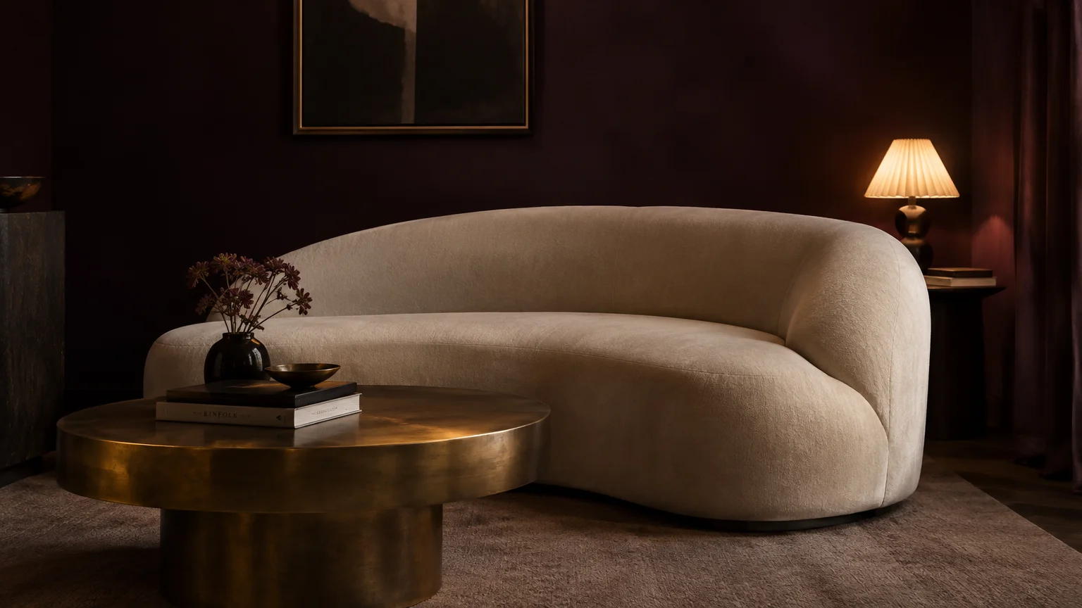 Curved cream sofa beside a round brass coffee table against dark moody aubergine walls with a lit table lamp.