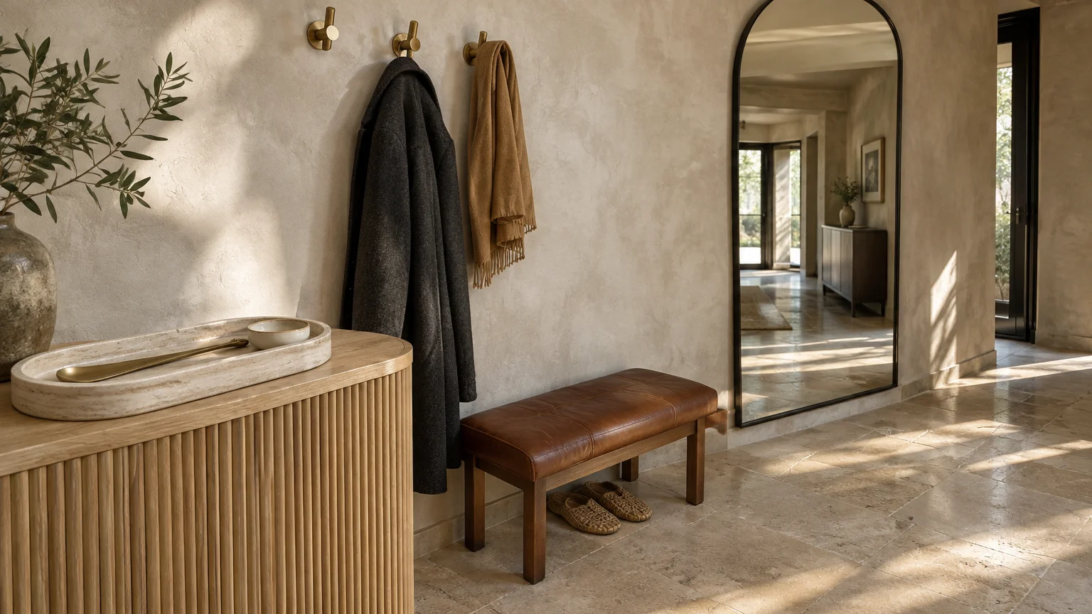 Foyer with a fluted wooden console, leather bench, woven slippers, arched floor mirror, and coats on wall hooks.