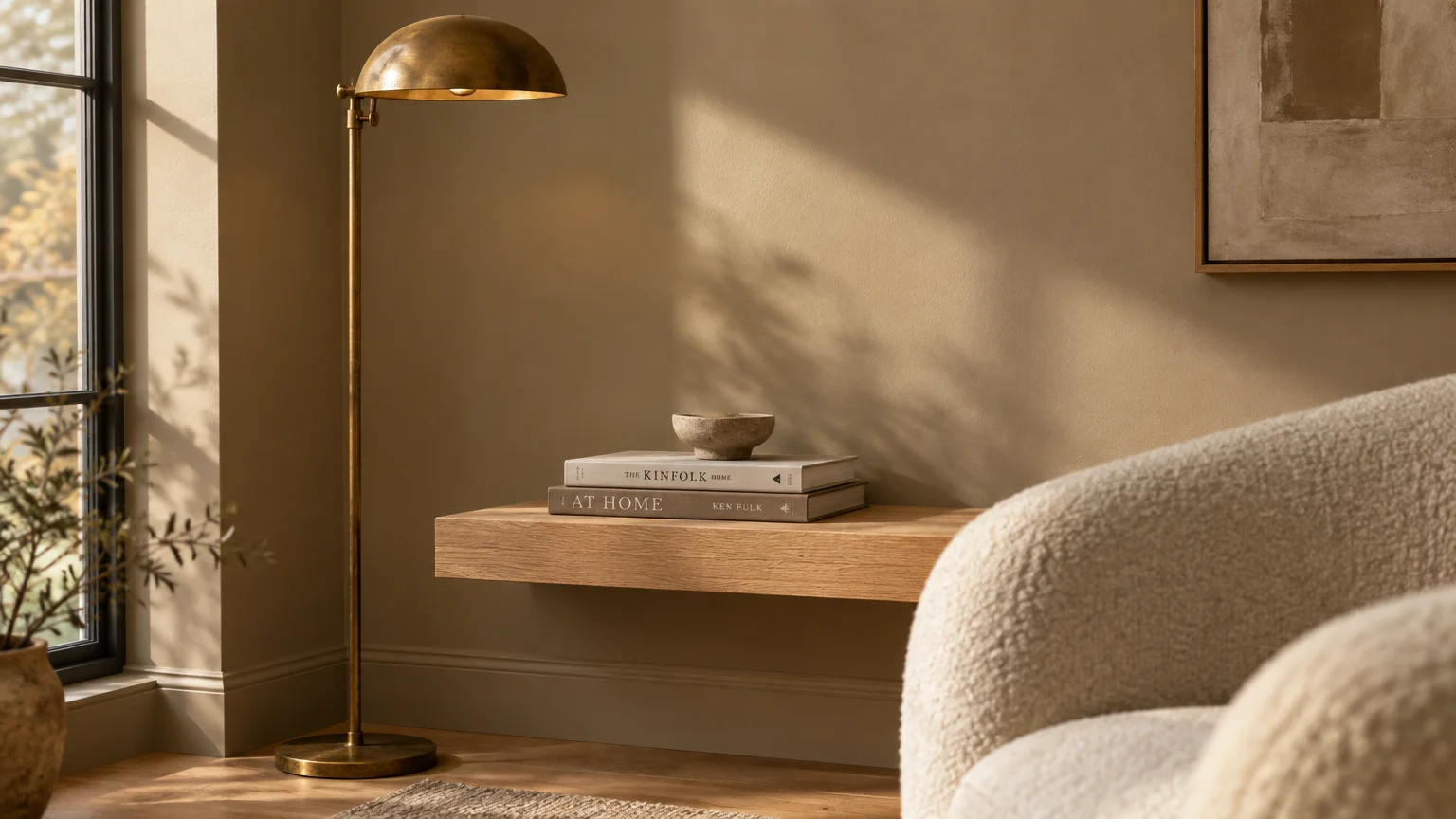 Brass floor lamp beside a floating wood shelf with stacked books, next to a beige wall and cream bouclé armchair.