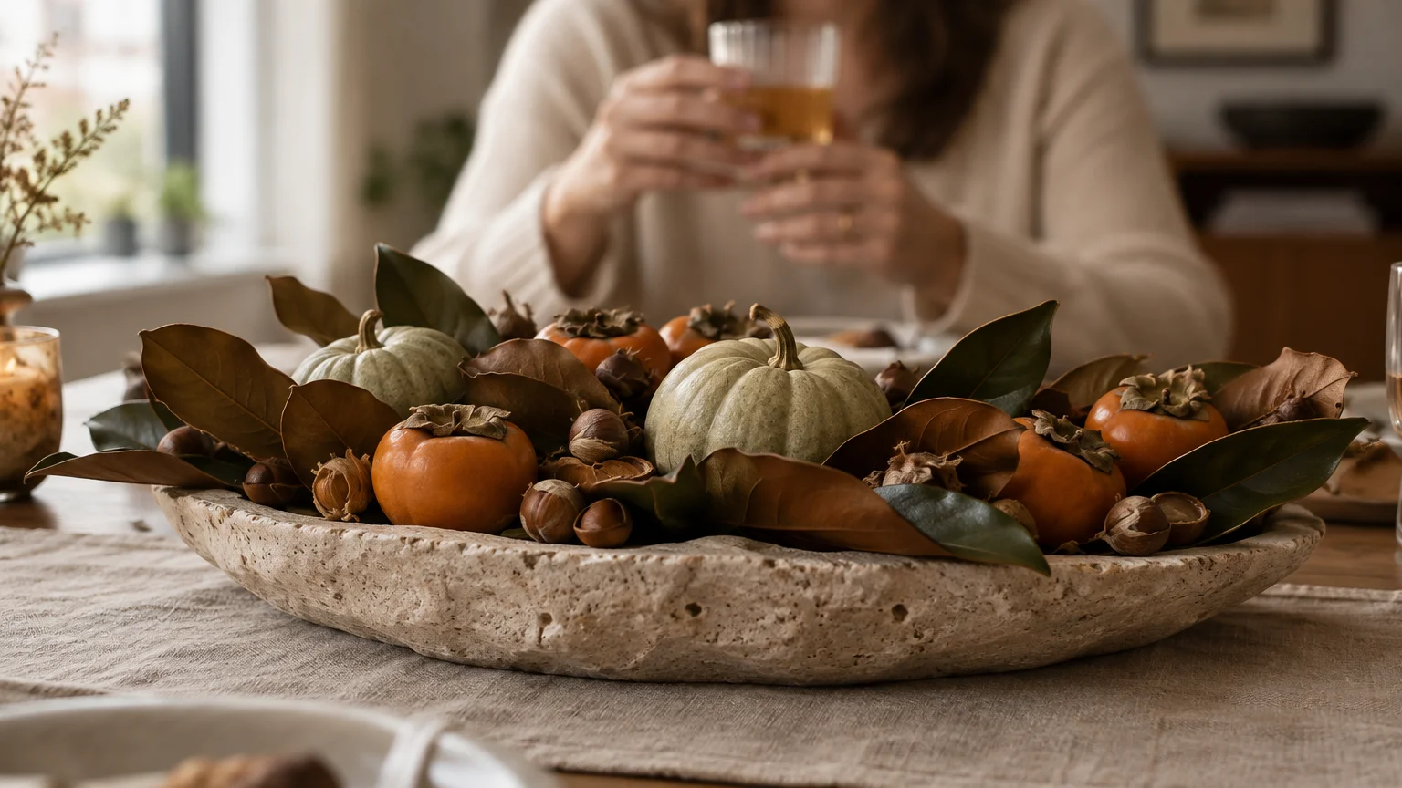 Shallow stone bowl filled with persimmons, small pale pumpkins, nuts, and magnolia leaves on a table before a seated guest.