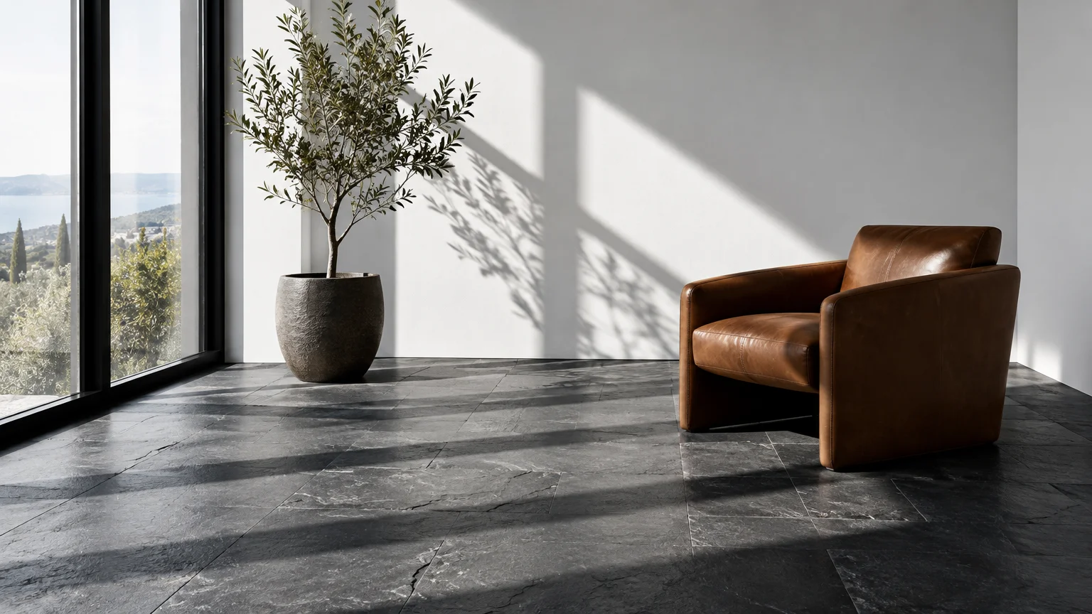 A brown leather armchair and potted olive tree sit on dark gray slate tile flooring against a sunlit white wall.