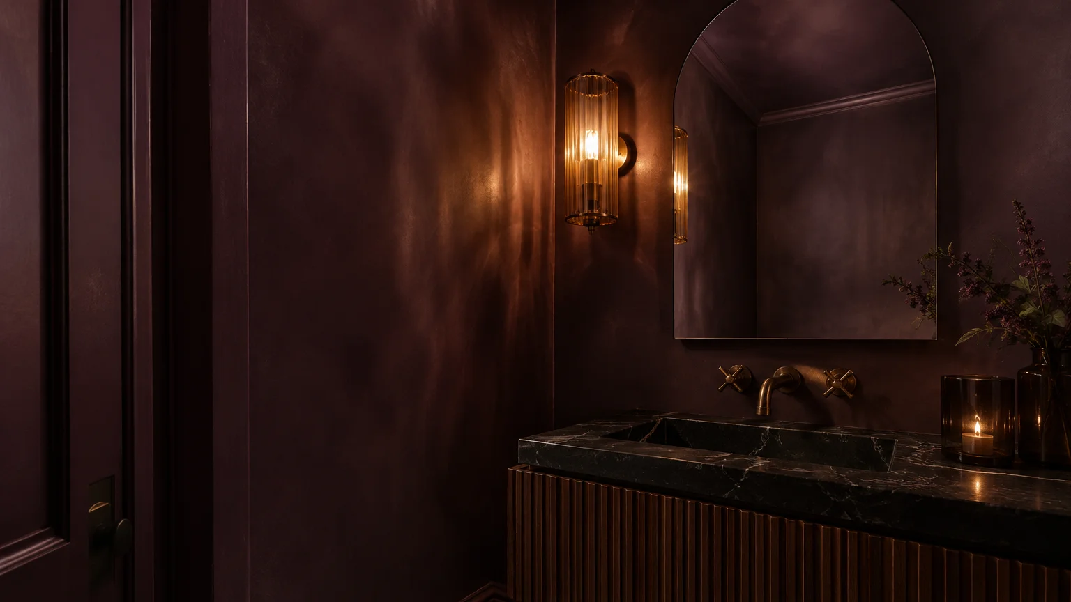Dark plum bathroom with an arched mirror, fluted vanity, black marble sink, and glowing wall sconce.