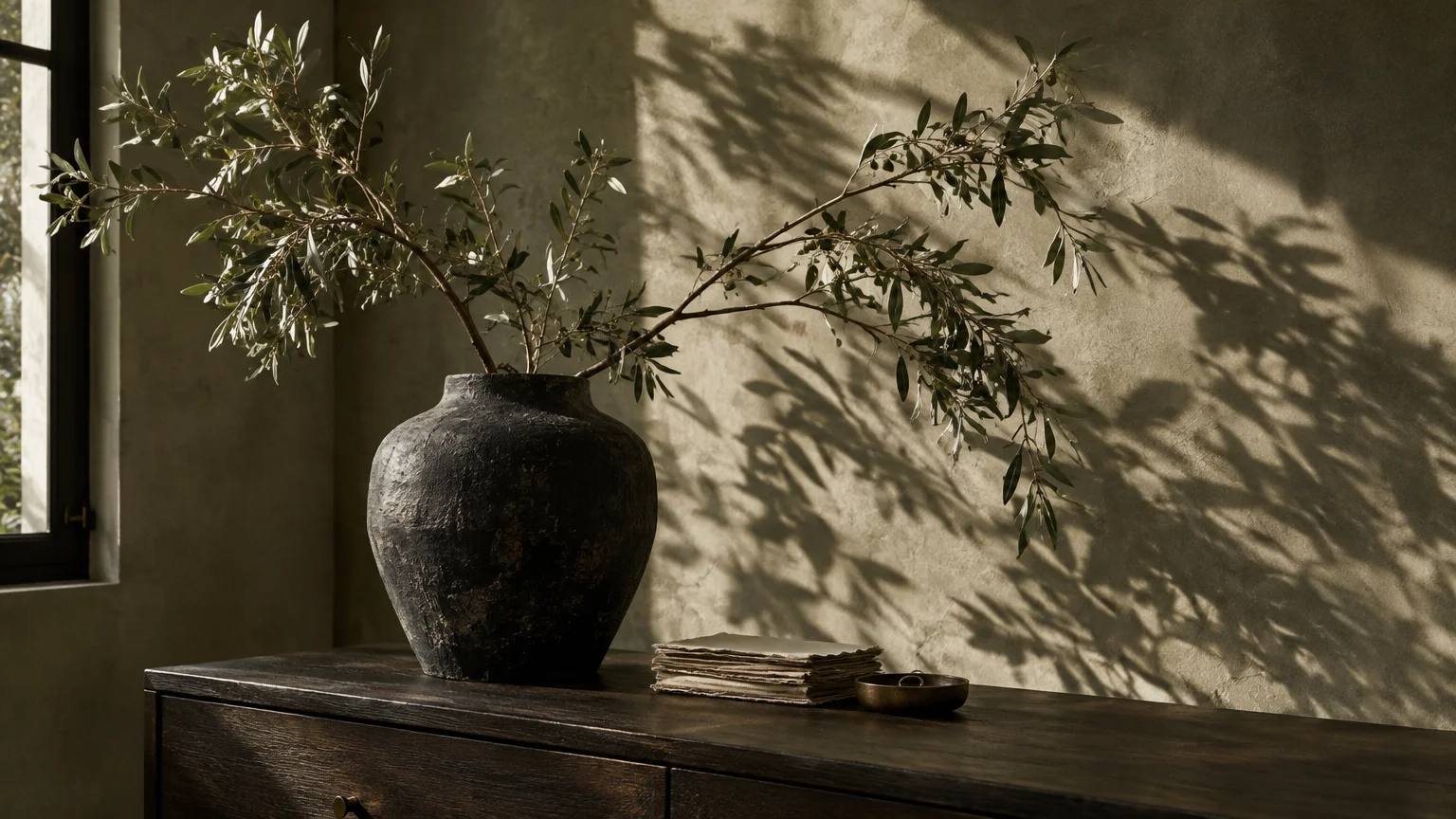 Green olive branches in a textured dark ceramic vase sitting on a dark wood dresser against a plaster wall.
