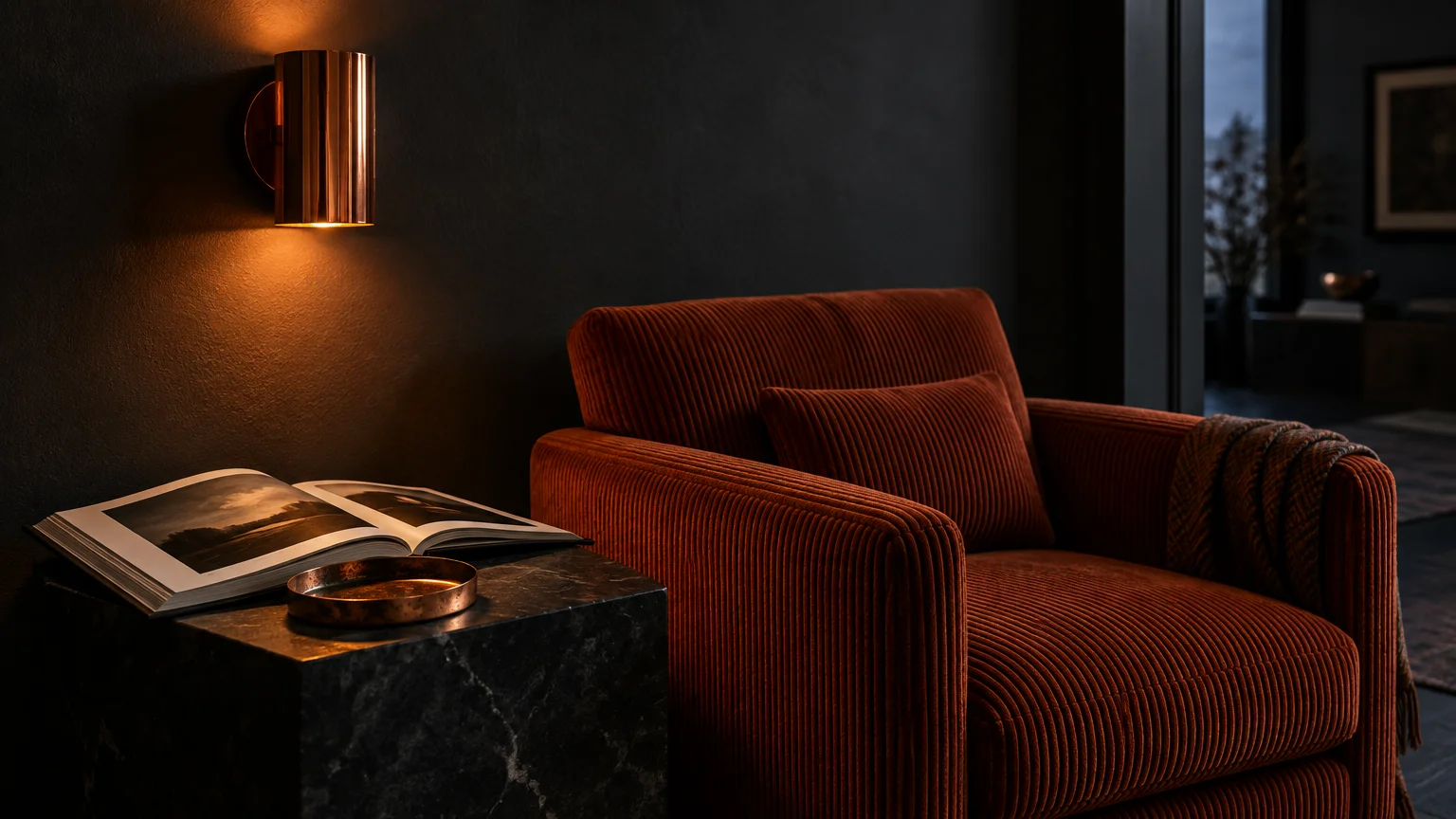 Rust corduroy armchair beside a dark marble side table and glowing copper wall sconce against charcoal walls.