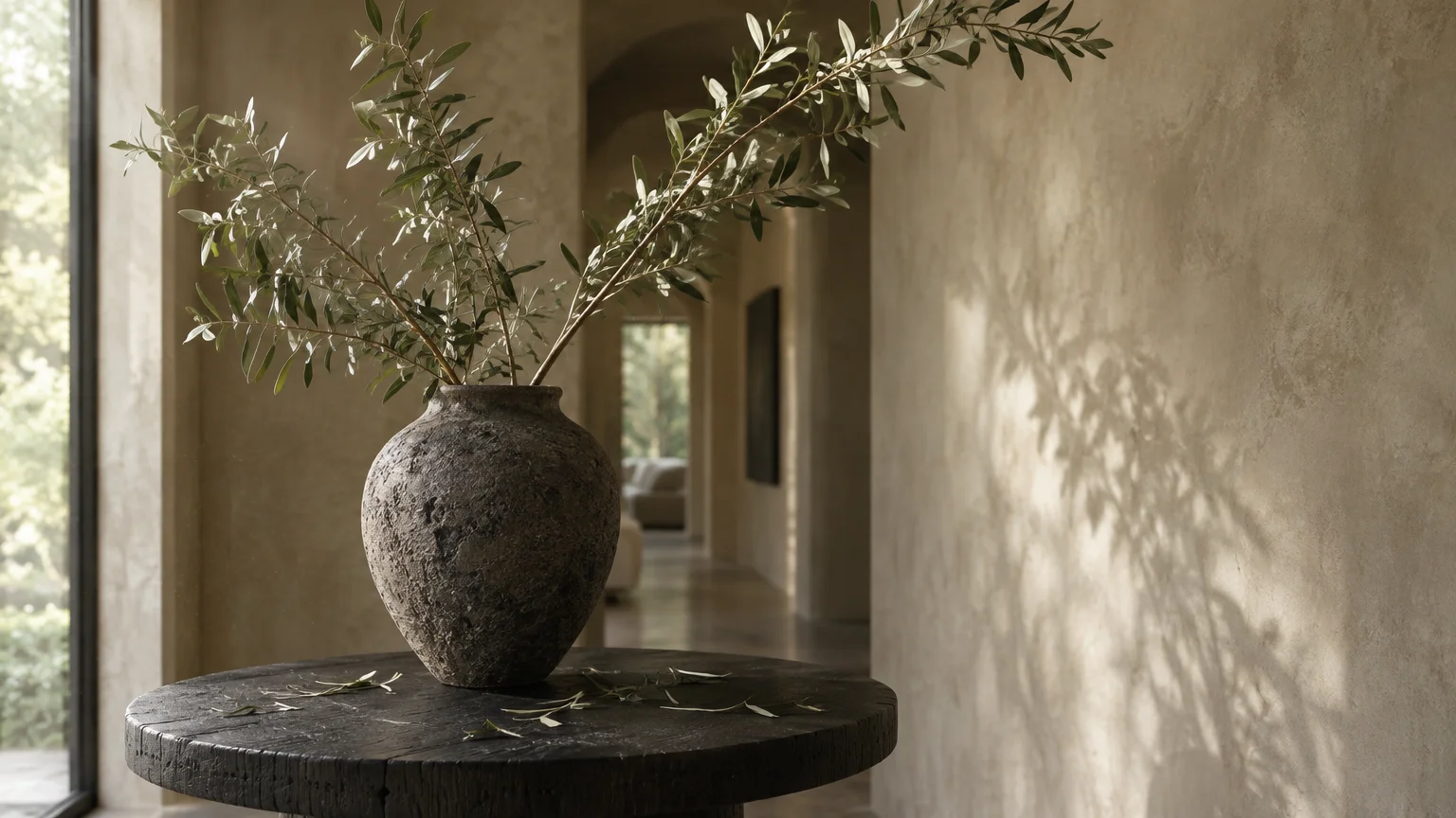 Rustic ceramic vase holding leafy green branches on a round wooden table in a hallway with textured plaster walls.