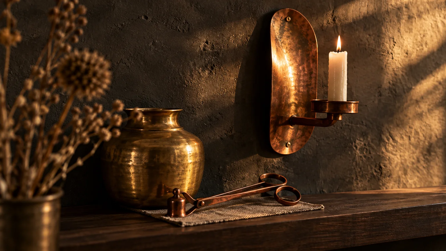 Lit candle in a copper wall sconce beside a brass pot and candle snuffer on a dark wooden shelf.