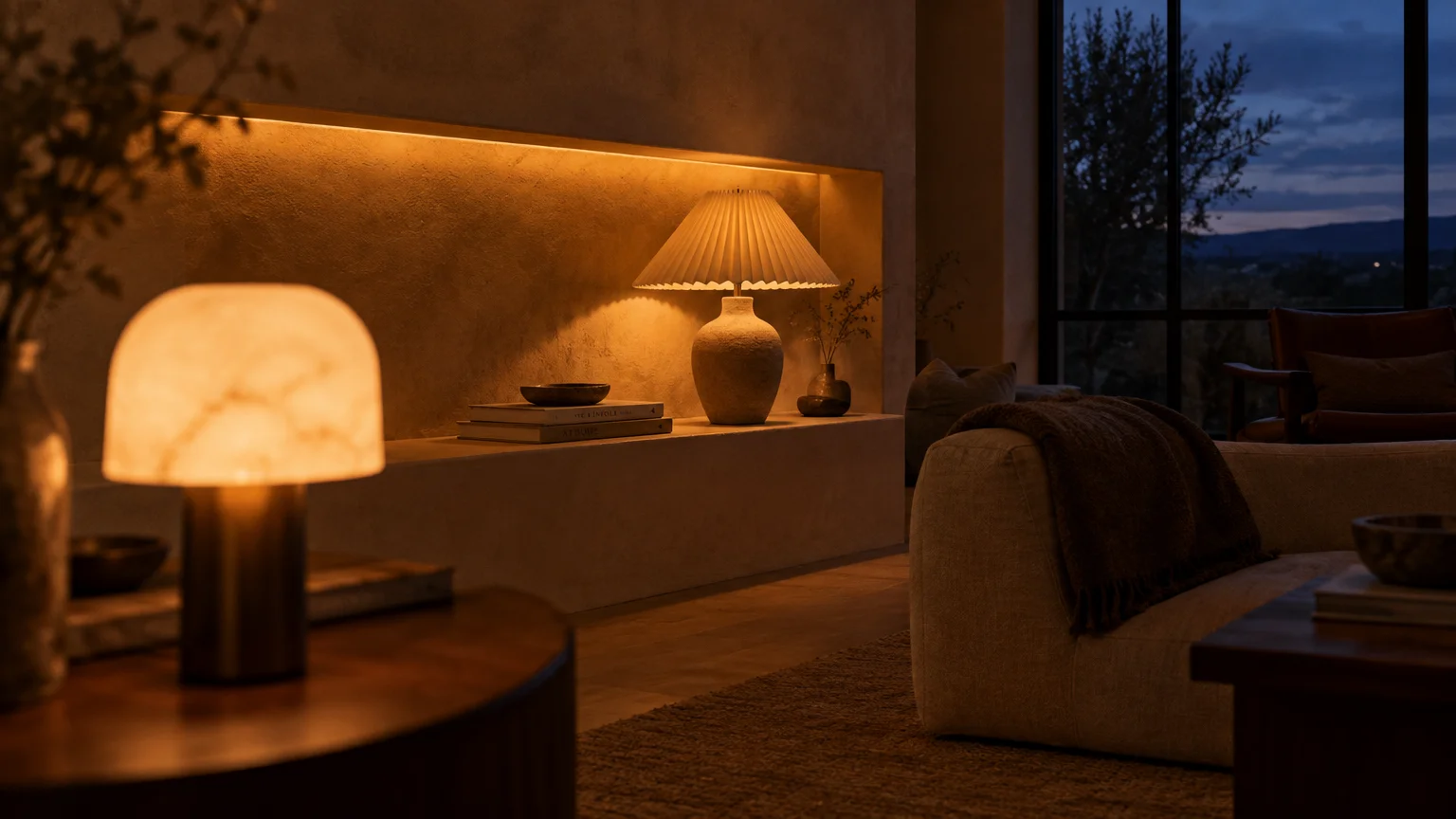 Dimmable table lamps and soft cove lighting cast a warm golden glow across a cozy living room at dusk.