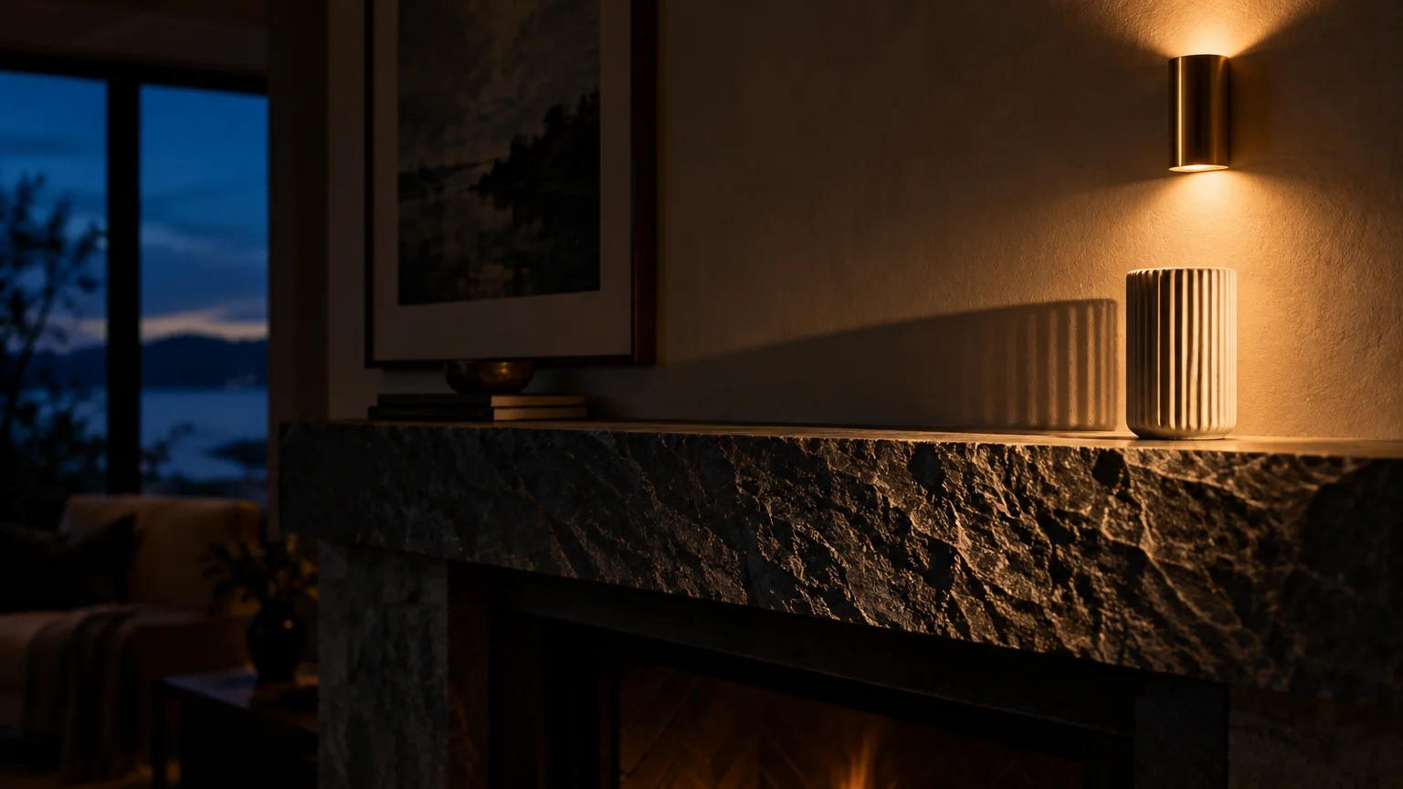 Modern stone fireplace mantel with a lit cylinder sconce casting dramatic shadows over a fluted ceramic vase.